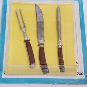 May include: A set of three miniature metal utensils, including a fork, knife, and spoon. The utensils are silver with brown plastic handles. The utensils are displayed in a clear plastic package with a yellow background.