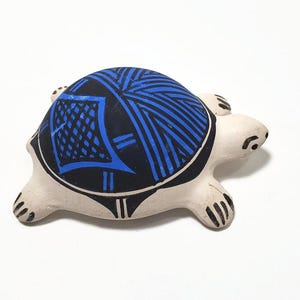 May include: A ceramic turtle figurine with a matte off-white body and a black shell featuring vibrant blue geometric line patterns. The turtle has four legs with black painted claws and a small head with simple black markings for eyes and a mouth.