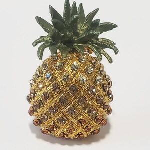 May include: A pineapple-shaped brooch featuring a gold-tone metal base with a textured, diamond-patterned surface. The body is encrusted with numerous clear and amber-colored rhinestones, while the top features a dark green metal leaf crown.