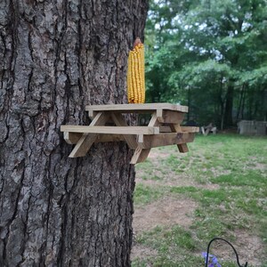 May include: A miniature wooden picnic table is attached to a tree trunk, with an ear of yellow corn on top. The table is made of light-colored wood and has a classic picnic table design. The background shows a grassy area and trees.