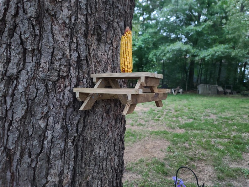 May include: A miniature wooden picnic table is attached to a tree trunk, with an ear of yellow corn on top. The table is made of light-colored wood and has a classic picnic table design. The background shows a grassy area and trees.