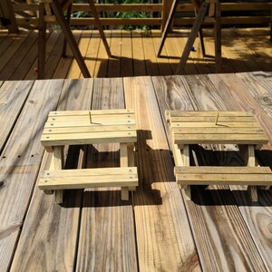May include: Two small, unfinished wooden picnic tables with slatted tops and attached benches sit on a wooden deck. The tables are made of light-colored wood and are designed for small items or decorative use.