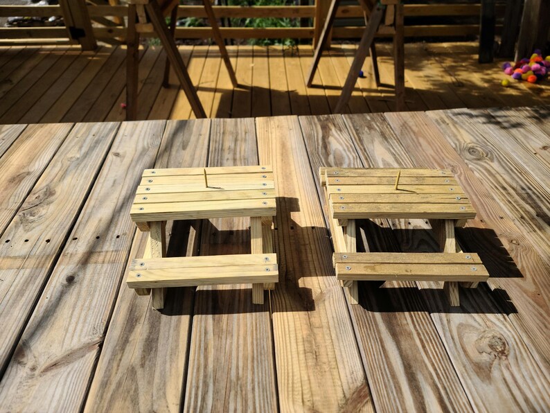 May include: Two small, unfinished wooden picnic tables with slatted tops and attached benches sit on a wooden deck. The tables are made of light-colored wood and are designed for small items or decorative use.