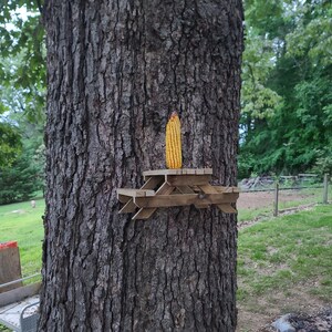 May include: A wooden miniature picnic table is attached to a tree trunk, with an ear of corn placed on top. The tree's bark is rough and brown. The background shows a grassy area and trees.