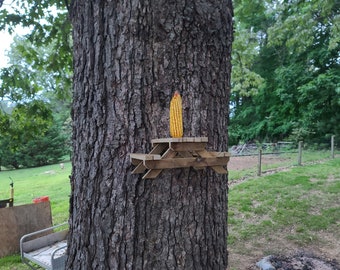 May include: A wooden miniature picnic table is attached to a tree trunk, with an ear of corn placed on top. The tree's bark is rough and brown. The background shows a grassy area and trees.