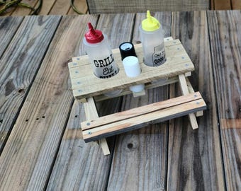 May include: A small wooden picnic table with two squeeze bottles and two small containers. One bottle reads "GRILL EAT" and the other reads "Taste so GOOD". The table is made of light-coloured wood. The bottles have red and yellow caps.