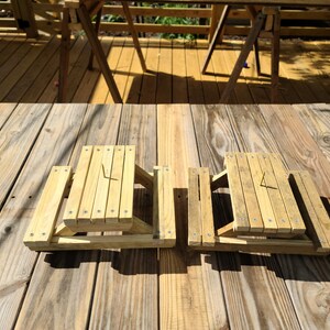 May include: Two small, unfinished wooden picnic tables with attached benches. The tables are constructed with vertical slats and secured with screws. The tables are sitting on a wooden deck.