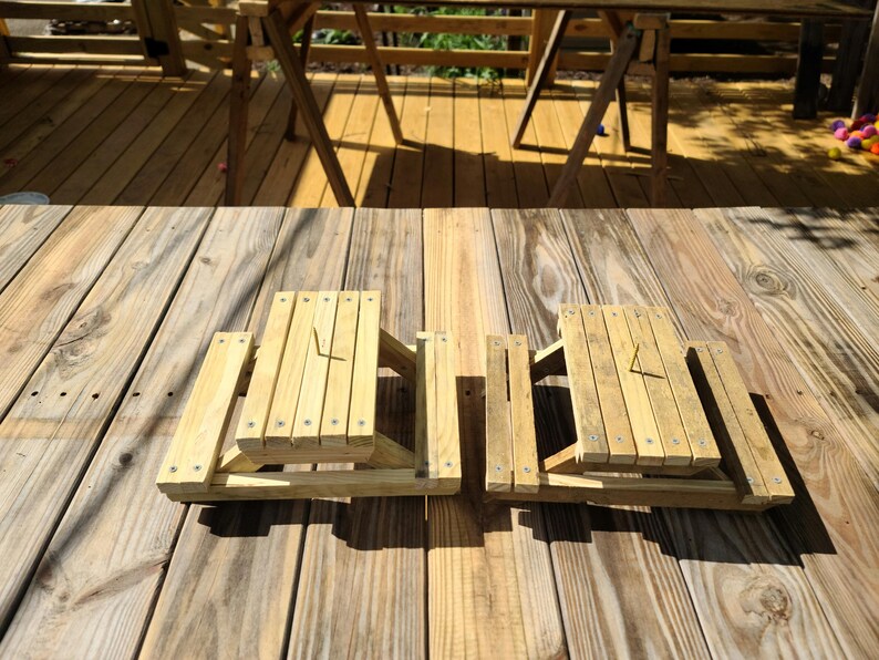 May include: Two small, unfinished wooden picnic tables with attached benches. The tables are constructed with vertical slats and secured with screws. The tables are sitting on a wooden deck.