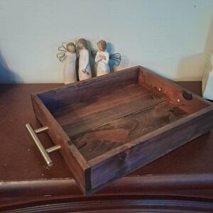 May include: A rectangular wooden tray with a dark brown finish and brass handles. The tray is empty and sits on a dark brown surface. Decorative figurines are in the background.
