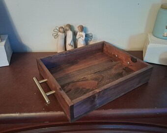 May include: A rectangular wooden tray with a dark brown finish and brass handles. The tray is empty and sits on a dark brown surface. Decorative figurines are in the background.
