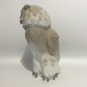 May include: A ceramic owl figurine in shades of beige and white. The owl is depicted in a side profile, with detailed feather textures and a slightly open beak. The figurine has a glossy finish and stands on a white surface.