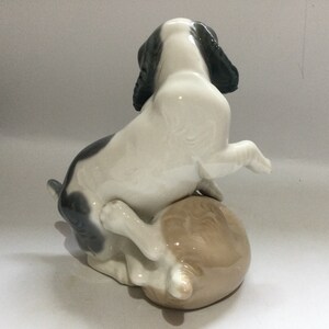 May include: A ceramic figurine of a dog in a playful pose, with its front paws raised. The dog has a white body with black and gray markings on its ears and back. It is perched on a light brown sphere.