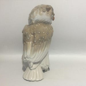 May include: A ceramic owl figurine in shades of beige, brown, and white. The owl is depicted in a seated position, with detailed feather textures and an orange beak. The figurine has a glossy finish and is likely a decorative item.