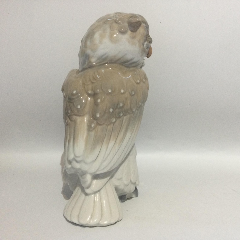 May include: A ceramic owl figurine in shades of beige, brown, and white. The owl is depicted in a seated position, with detailed feather textures and an orange beak. The figurine has a glossy finish and is likely a decorative item.