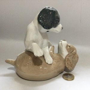 May include: A ceramic figurine depicting two puppies at play. One puppy is white with black markings, sitting on top of a light brown puppy. The figurine is glossy and detailed, showcasing a playful scene. A coin is next to the figurine.