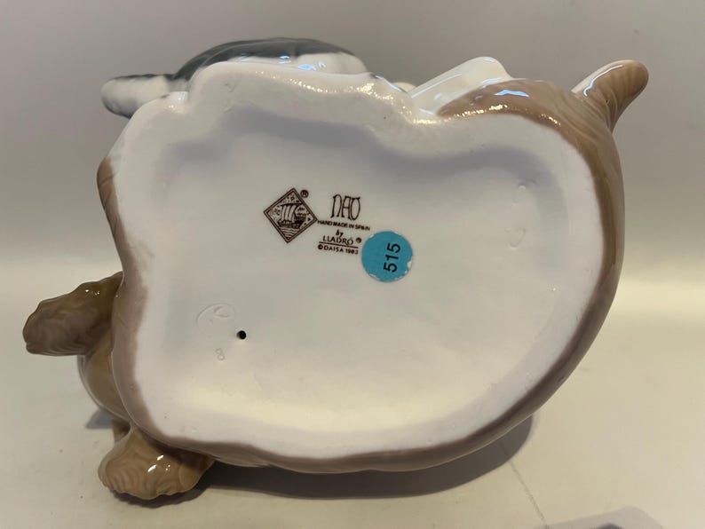 May include: A ceramic figurine of a seated animal, possibly a rabbit or dog, with a white belly and brown and gray accents. The figurine has a glossy finish and a blue sticker with the number 515.