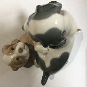 May include: A ceramic figurine depicting two dogs in a playful embrace. One dog is light brown and white, while the other is primarily white with gray patches. The glossy finish enhances the details of the dogs' features.