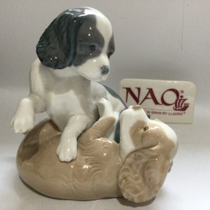 May include: A porcelain figurine of two playful puppies. One puppy is light brown and white, lying on its back. The other puppy is white, black, and gray, standing above. The figurine has a glossy finish. A rectangular sign with the text "NAO" is in the background.