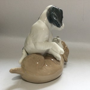 May include: A ceramic figurine depicting two playful dogs. One dog is white with black markings, playfully positioned atop a light brown dog. The figurine has a glossy finish and is set against a plain background.