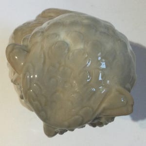 May include: A light beige ceramic figurine of a stylized animal, possibly a bird or owl. The sculpture has a textured surface with a glossy finish. The figurine has a rounded body and a prominent beak.