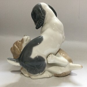 May include: A ceramic figurine depicting two dogs in a playful pose. One dog is white with black markings, the other is tan. The dogs are interacting, with one sitting on top of the other. The figurine has a glossy finish.