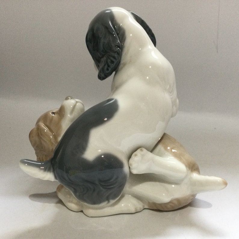 May include: A ceramic figurine depicting two dogs in a playful pose. One dog is white with black markings, the other is tan. The dogs are interacting, with one sitting on top of the other. The figurine has a glossy finish.