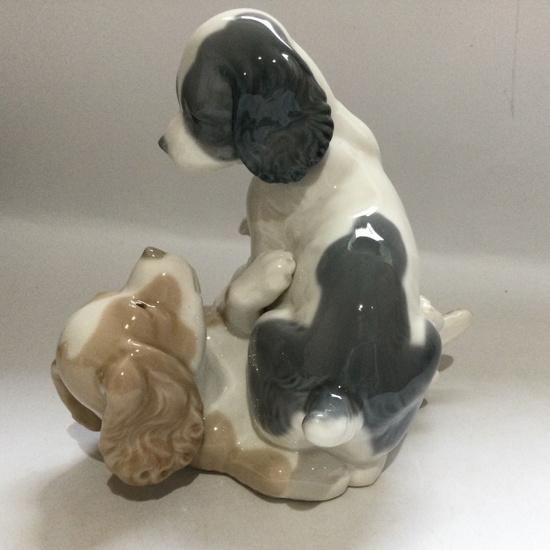 May include: A ceramic figurine depicting two playful puppies. One puppy is lying on its back, while the other is perched on top. The puppies are primarily white with patches of gray and tan. The figurine has a glossy finish.
