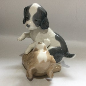 May include: A porcelain figurine depicting two playful puppies. One puppy is seated, with a black and white coat, while the other is lying down with a tan and white coat. The figurine is detailed and has a glossy finish.