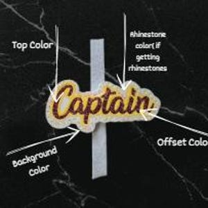 Op de afbeelding: Een op maat gemaakte woorduitsnede met de tekst Captain in een donkere glitterafwerking, voorzien van een lichte omlijning en achtergrondlaag. Het ontwerp bevat witte pijlen en labels die wijzen naar Top Color, Rhinestone color, Offset Color en Background Color.