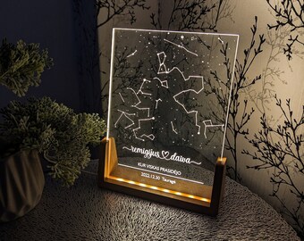 Op de afbeelding: Een verlicht acryl paneel toont een sterrenbeeldkaart met de tekst "remigijus ♡ daiva", "KUR VISKAS PRASIDĖJO" en de datum "2022.12.30 Tauragė". Het paneel staat in een houten voet met warmwitte LED-verlichting, wat een zachte gloed creëert.