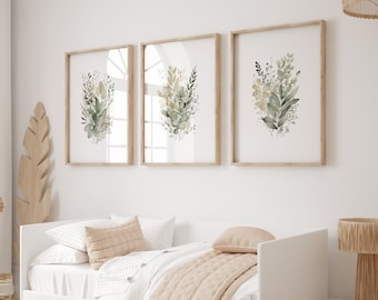 Op de afbeelding: Drie ingelijste aquarelprints van groen en geel gebladerte, hangend boven een wit dagbed met een beige deken en kussens.