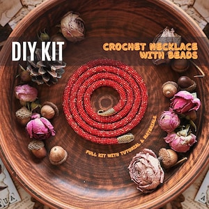May include: DIY kit for a crochet necklace with beads. The kit includes red beads, gold-colored clasps, and a tutorial. The necklace is coiled in a bowl with dried flowers and acorns.
