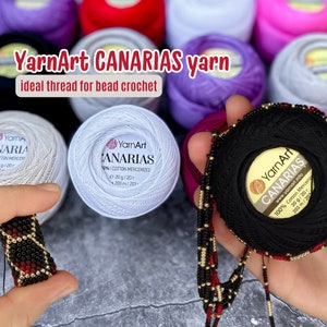 Può includere: Gomitoli di filato YarnArt Canarias in bianco, nero e rosso, un filo 100% cotone mercerizzato. L'etichetta riporta "YarnArt Canarias 100% Cotone Mercerizzato 20g/20г 203 m/203". Un girocollo di perline è in primo piano.