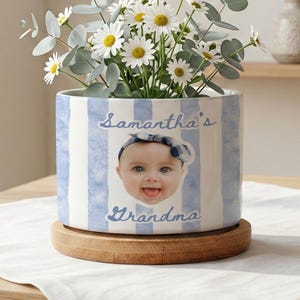 Könnte beinhalten: Keramik-Blumentopf mit blau-weißen Streifen, gefüllt mit Gänseblümchen und Eukalyptus. Der Topf zeigt ein Babyfoto und den Text "Samantha's Grandma". Er steht auf einem Holzsockel, ideal für eine Zimmerpflanze.
