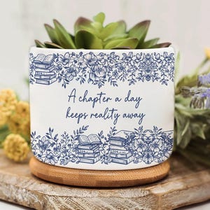 Op de afbeelding: Witte keramische plantenpot met een vetplant. De pot heeft een blauw bloemen- en boekontwerp met de tekst "A chapter a day keeps reality away". Hij staat op een ronde houten basis.