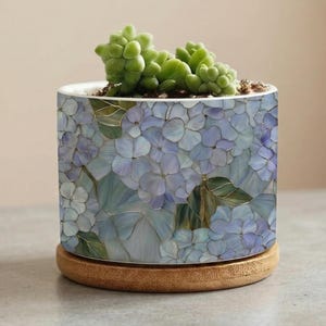 以下が含まれることがあります： 青いアジサイの花柄が施された円筒形のセラミック植木鉢。植木鉢は、丸い薄茶色の木製コースターの上に置かれています。緑色の多肉植物が植木鉢に植えられています。