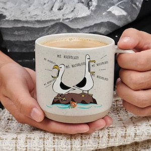 Op de afbeelding: Een beige gespikkelde keramische mok met een cartoonillustratie van twee zeemeeuwen op een rots, met de tekst "MS. NICHOLAS!". De mok is gevuld met een lichtbruine vloeistof en heeft een wit handvat.
