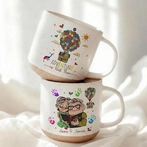 May include: Two white ceramic mugs stacked on top of each other. The top mug has a colorful illustration of a house floating with balloons, a sun, and the text "Adventure is Out There." The bottom mug features an illustration of an elderly couple and the text "Anna & James."