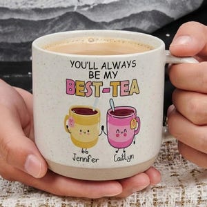May include: A speckled ceramic mug with the text "YOU'LL ALWAYS BE MY BEST-TEA" and two cartoon tea cups. One cup is yellow with the name "Jennifer", and the other is pink with the name "Caitlyn". The mug is filled with coffee.