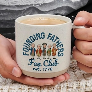 May include: A speckled ceramic coffee mug with the words "FOUNDING FATHERS Fan Club est. 1776" in blue. The mug features a cartoon illustration of six men in colonial attire. The mug is filled with a light brown liquid.