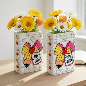 Puede incluir: Dos jarrones rectangulares blancos con un diseño de lápiz y manzana, llenos de flores amarillas, blancas y naranjas. Los jarrones tienen un lazo y el texto "Ms. Lisa". Los jarrones están sobre una superficie de madera.