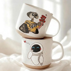 May include: Two white ceramic mugs stacked on top of each other. The top mug features a yellow robot holding flowers, and the bottom mug has a white robot with a heart above it. Both mugs have brown bottoms and white handles.