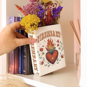 May include: A white ceramic vase with the word "FIREHEART" and a heart with flames design, filled with colorful dried flowers. The vase is rectangular and sits on a white shelf next to a row of books.