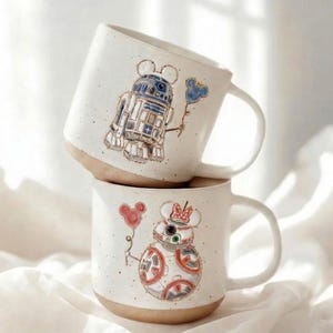 May include: Two stacked white ceramic mugs with brown bases. The top mug features a blue and silver R2-D2 design with a blue balloon. The bottom mug has a BB-8 design with a red bow and a red heart balloon. Both mugs have a speckled finish.