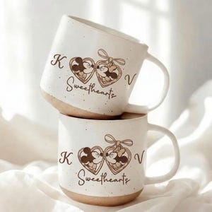 Può includere: Due tazze in ceramica bianca impilate, con base marrone. Ogni tazza presenta un design a forma di cuore con Topolino e Minni, la parola "Sweethearts" e le lettere "K" e "V".