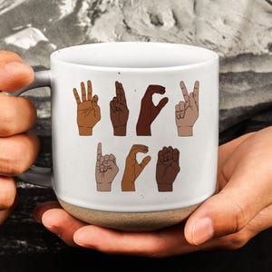 May include: White ceramic mug with a gray handle and a tan base. The mug features illustrations of hands spelling out the word "LOVE" in American Sign Language. The hands are depicted in various skin tones.