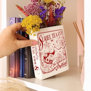May include: A white rectangular vase with the text "SURIEL TEA CO" in red, filled with colorful dried flowers. The vase has a decorative design featuring a teacup, teapot, and floral elements. The text "The hottest tea in Prythian" is also visible.