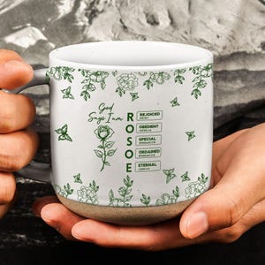 Op de afbeelding: Witte keramische mok met een bloemenontwerp en de tekst "God Says I Am ROSE". De mok heeft groene bloemenillustraties en tekst, met een natuurlijk gekleurde basis. De mok wordt door een persoon vastgehouden.