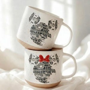 May include: Two white ceramic mugs stacked on top of each other. Each mug features a black graphic design of the Death Star and TIE fighters, with a red bow on the top mug. The mugs have a speckled finish and a tan base.