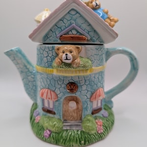 Puede incluir: Una caprichosa tetera de cerámica con forma de casa, con un exterior azul claro con un patrón de piedra. La tetera tiene un tejado rosa, una puerta marrón y está adornada con figuras de osos de peluche. La base está decorada con hierba verde y flores.